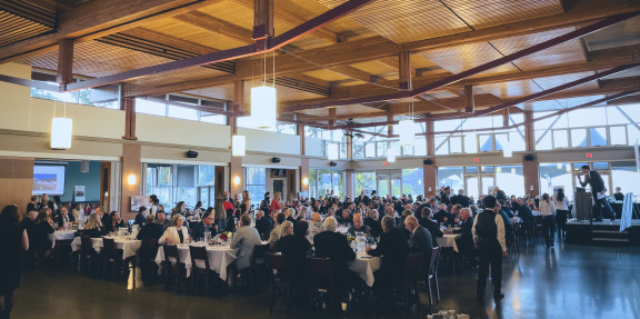 A large crowd sits around linen-dressed tables enjoying their meal, with floor to ceiling windows and ocean views in background