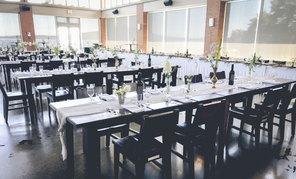 Brightly lit by the sun (floor to ceiling windows front and right), we can see tables set for dinner with a fully draped high table beyond