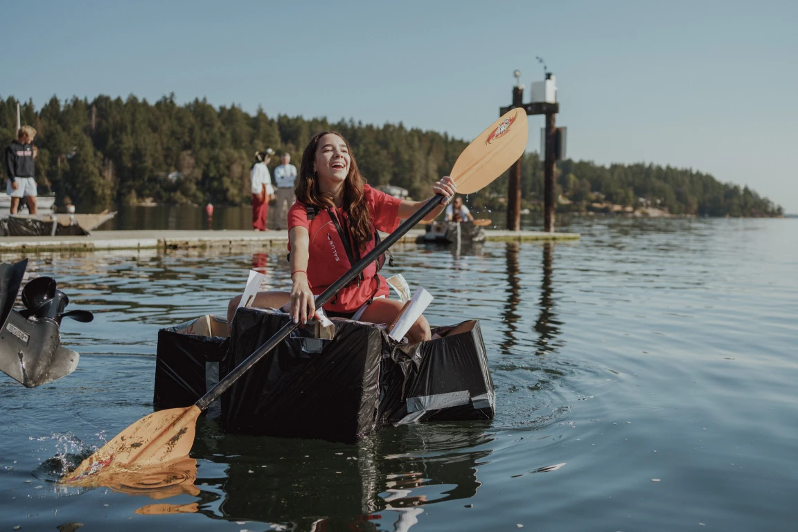 A student laughing while paddling a homemade cardboard boat on the water