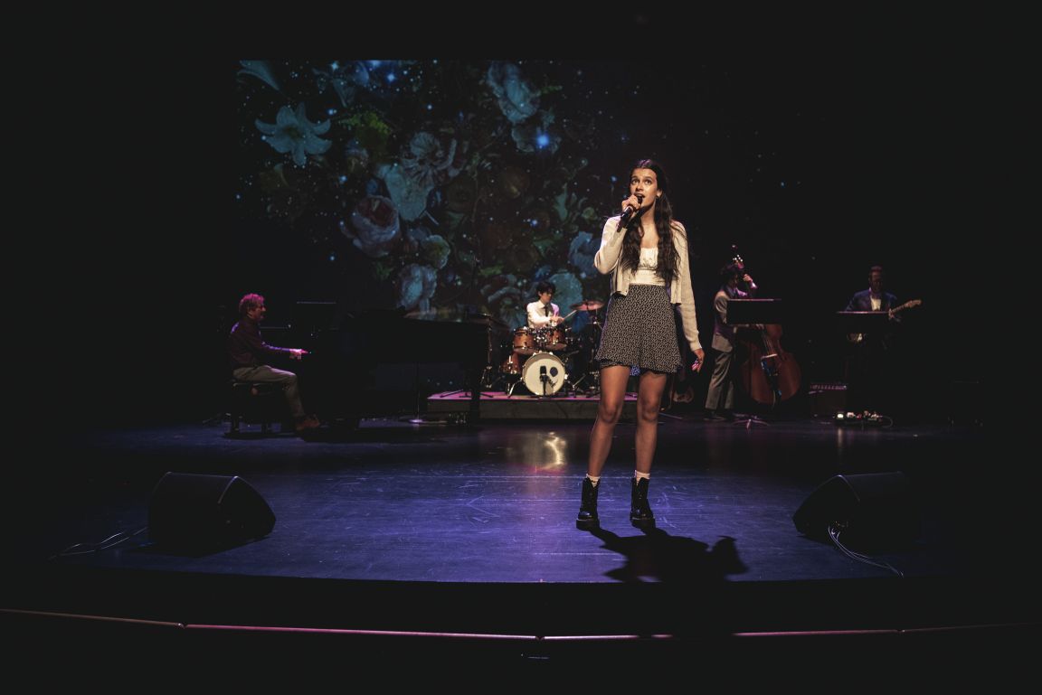A student singing on stage with a band, seen from the theatre floor