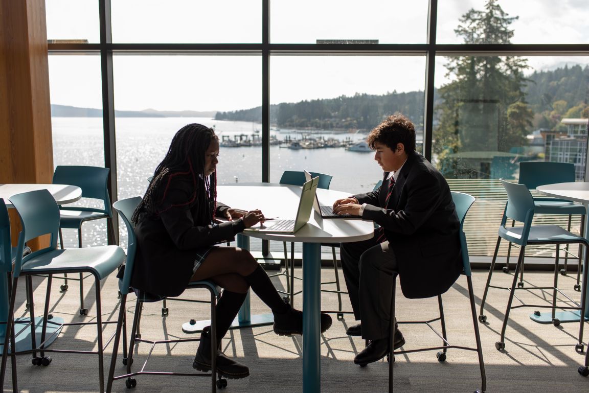 Two students working at a laptop by the window overlooking the water