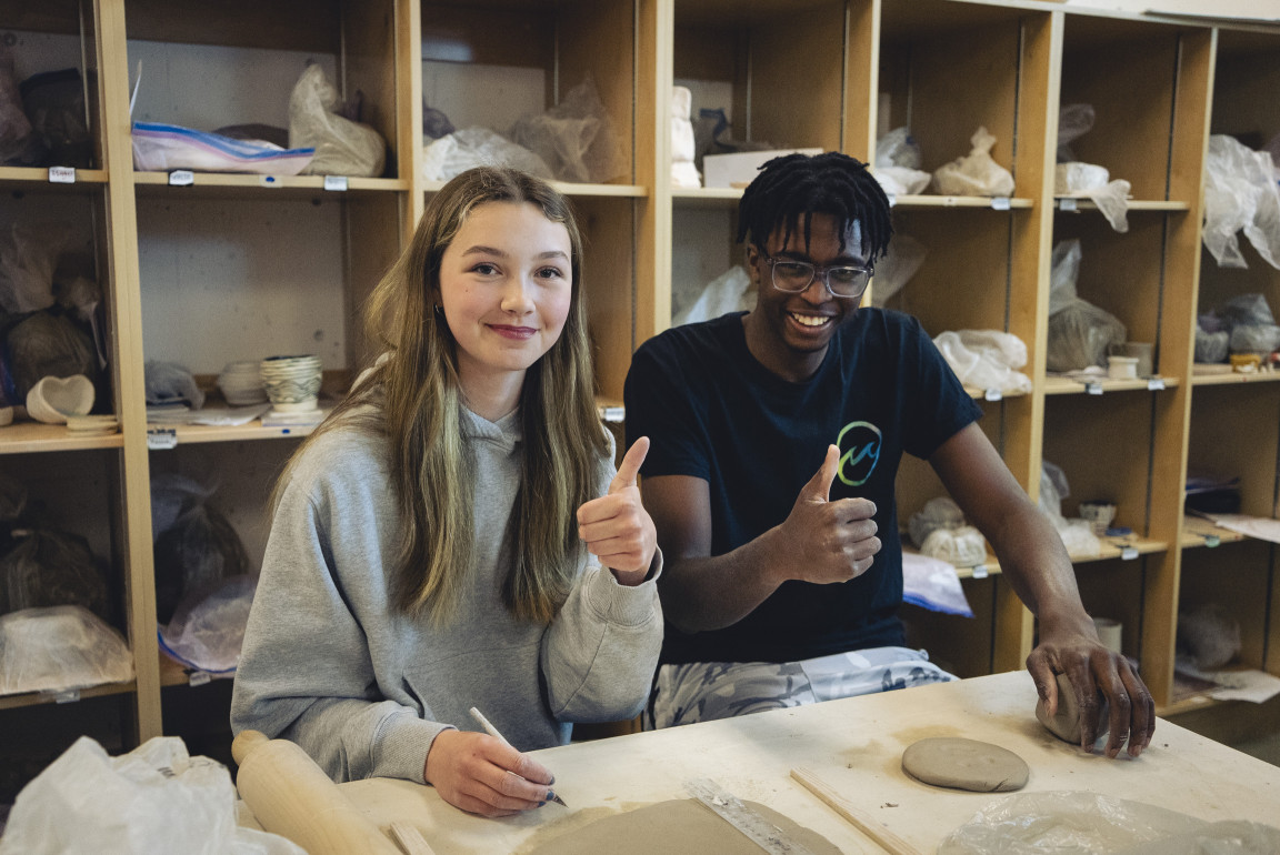 Two ceramics students giving a thumbs up at their hand-building table