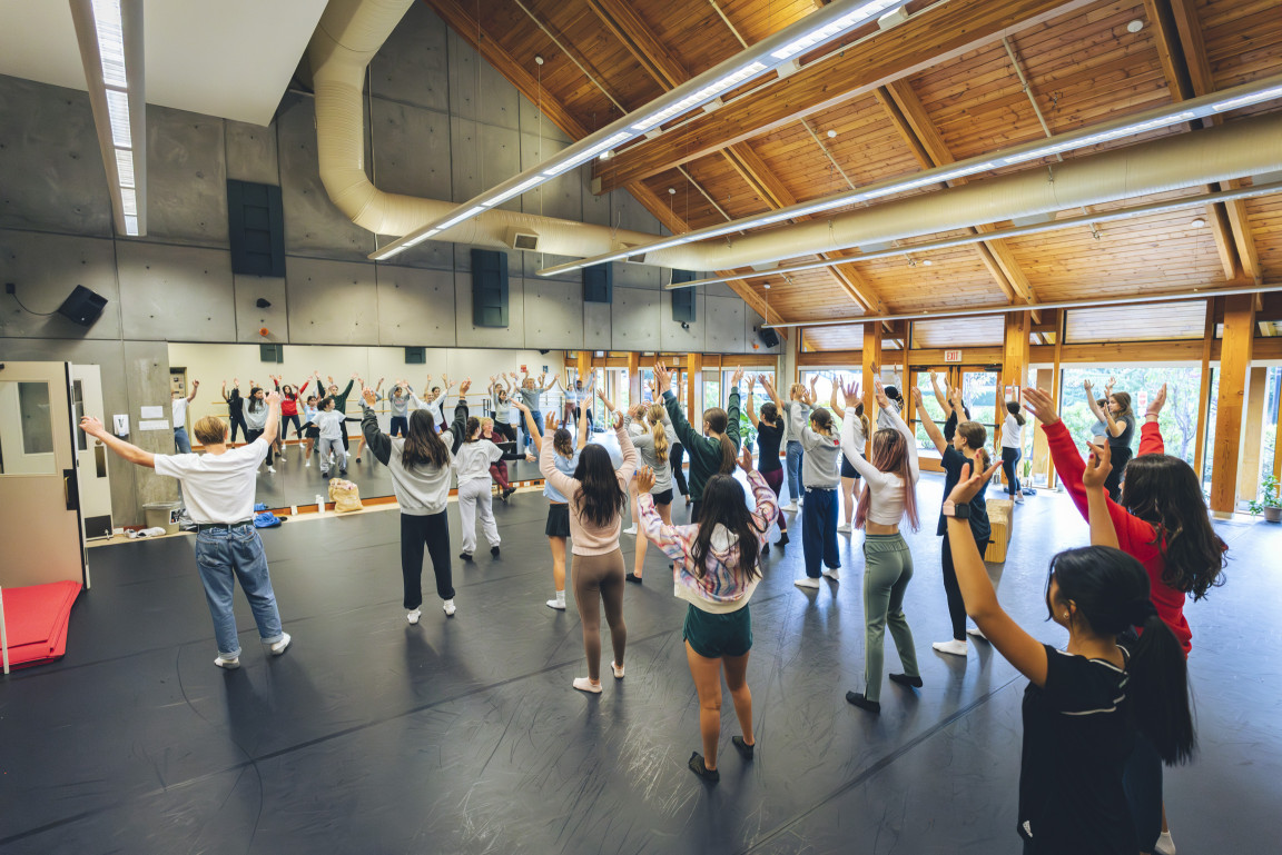 A large dance class rehearsing with arms raised in the studio