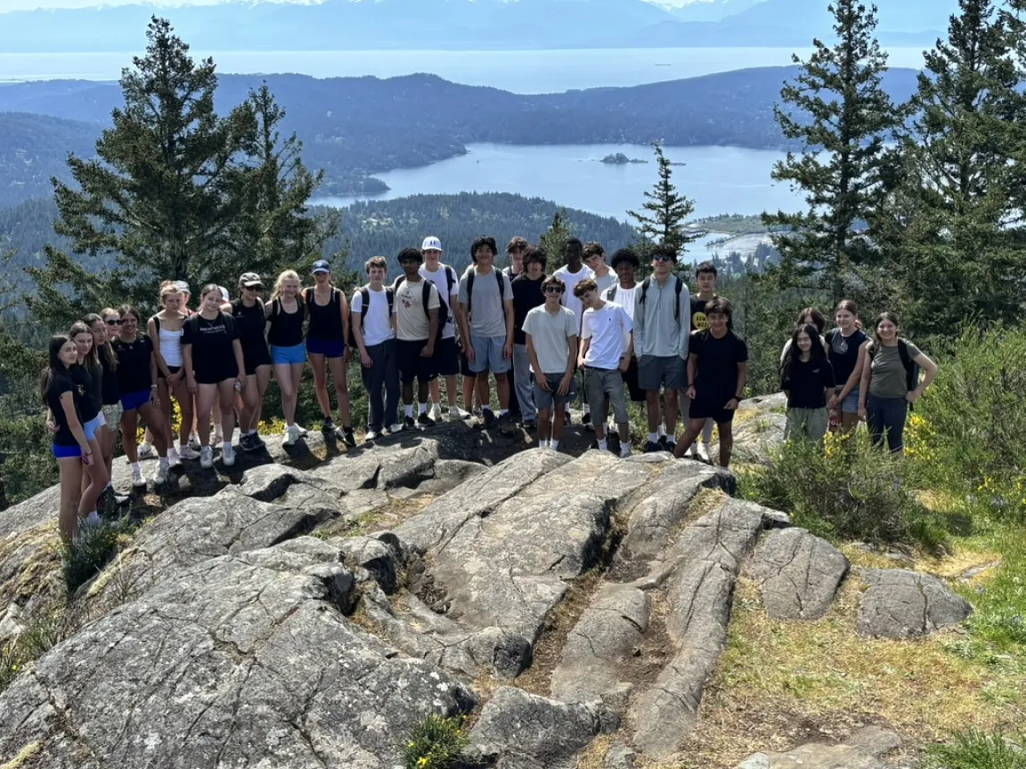 A large group of youth posing on a rocky peak overlooking a scenic coastal landscape with islands