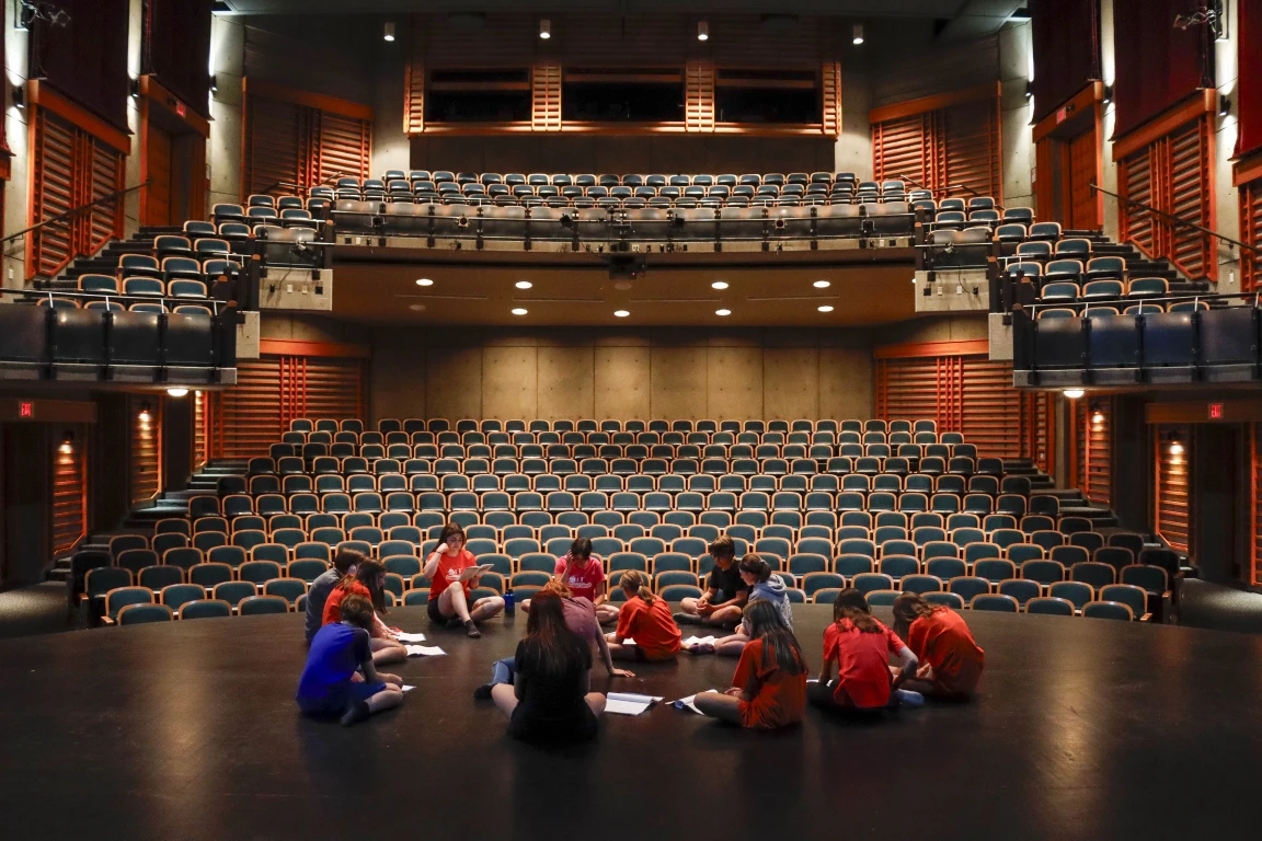 Young children sit cross-legged on a stage studying a script