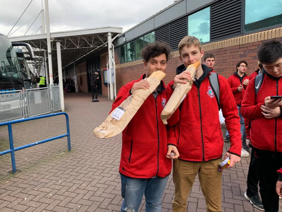 Two students in red jackets posing with long baguettes outside a bus station
