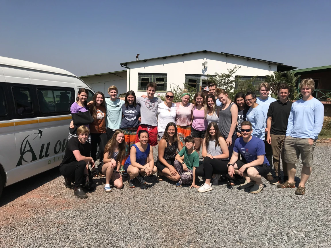 A large group of people posing for a photo in front of an All Out Africa van and a white building