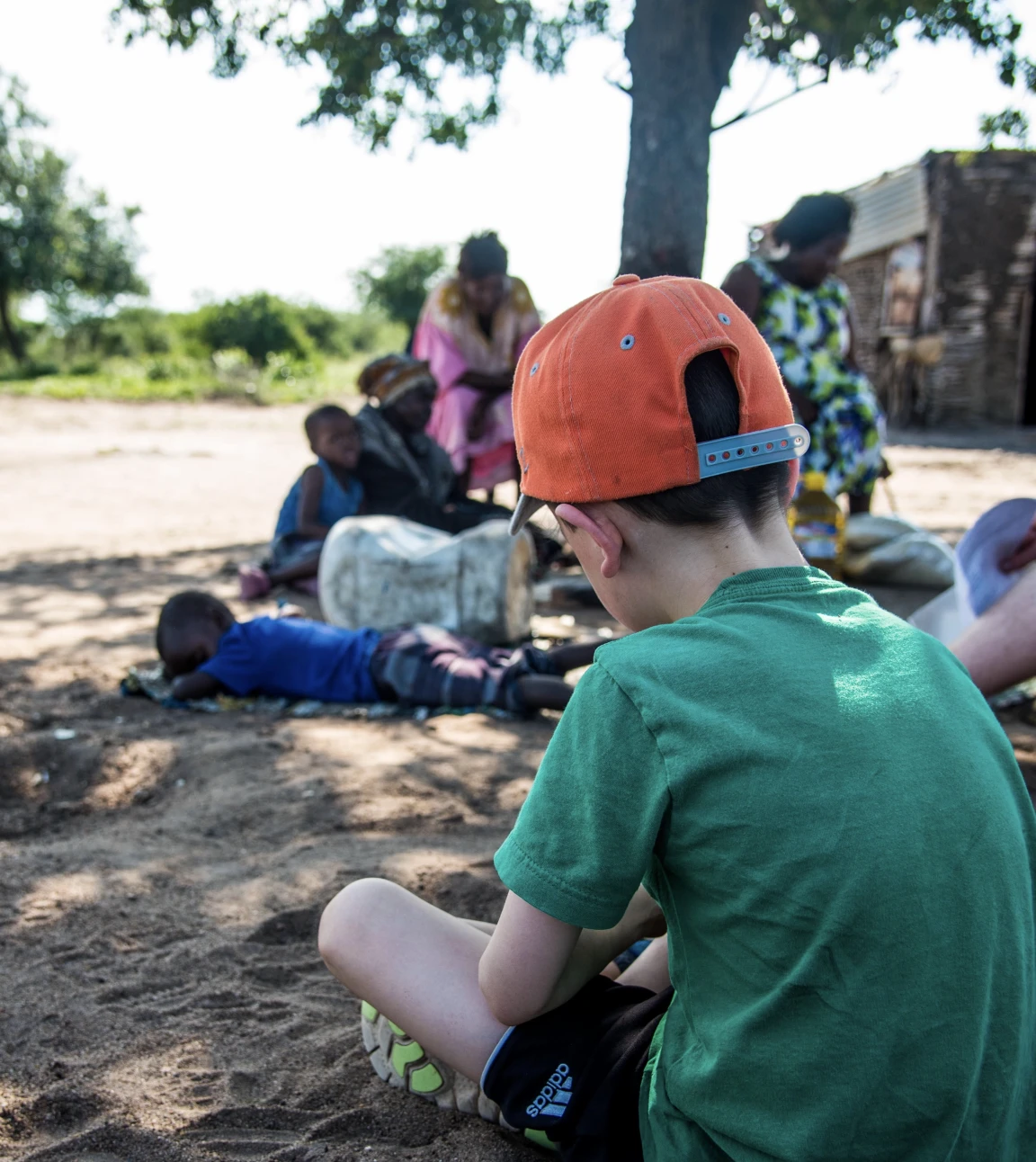 A boy in an orange hat and green shirt sits on the ground, looking toward a group of people under a tree