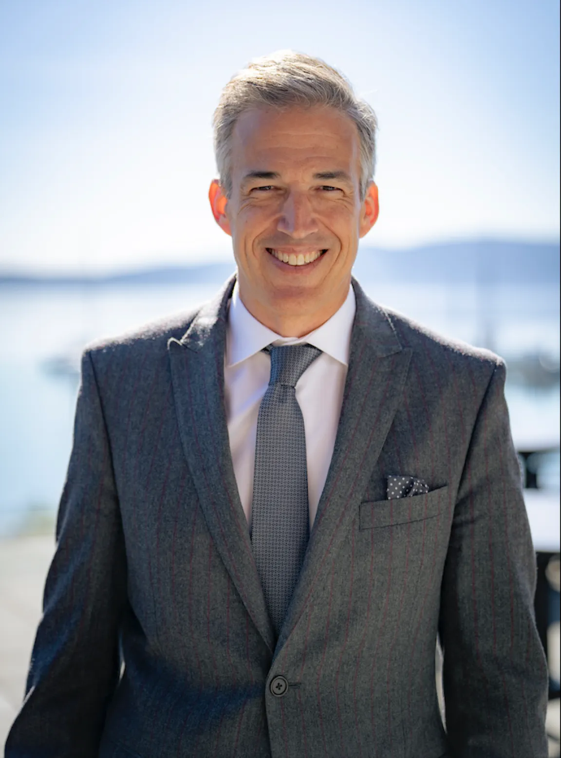 A man in a grey suit smiling by the waterfront