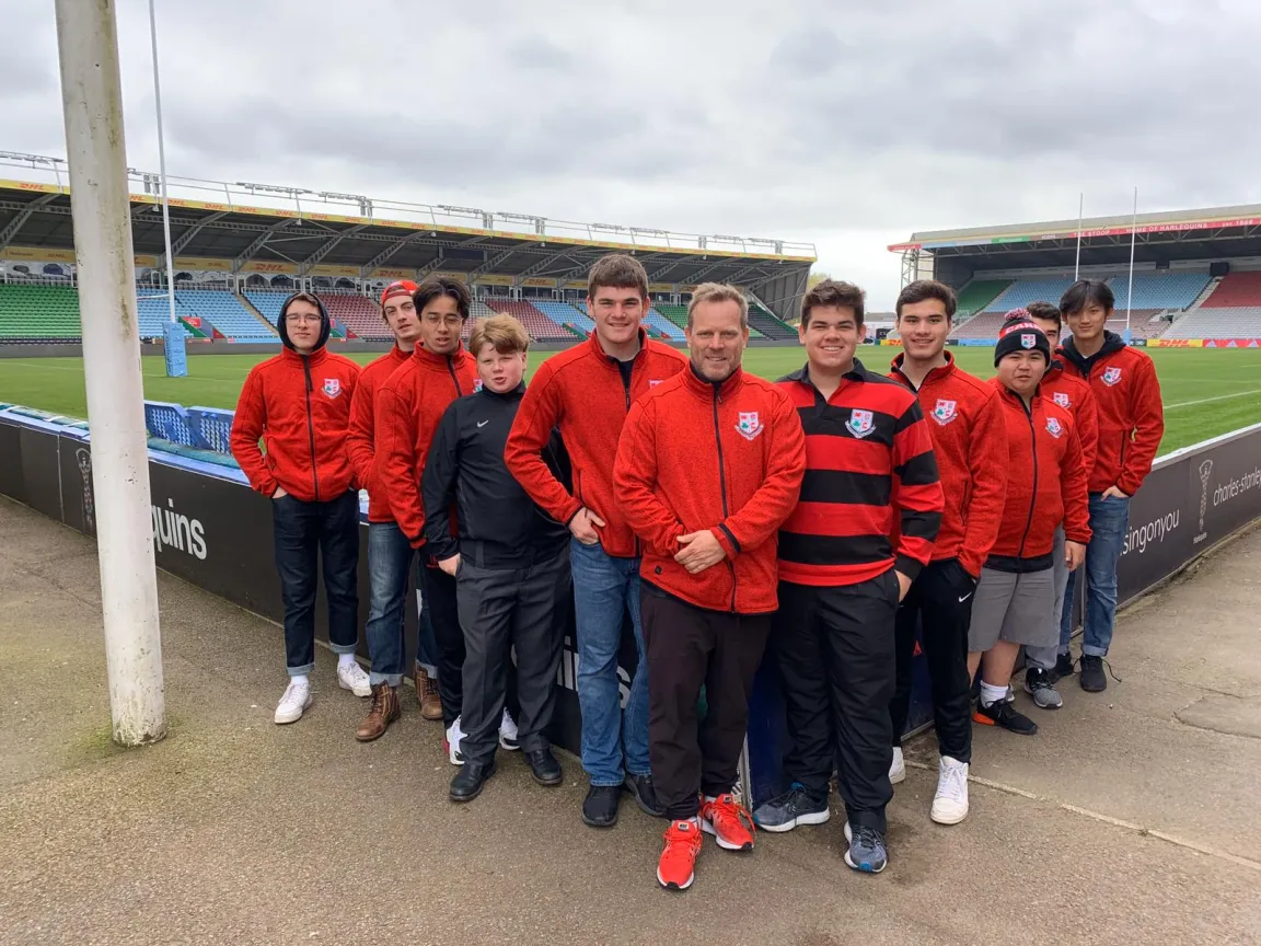 A group of young men and an adult in red team jackets posing on the sidelines of a rugby stadium